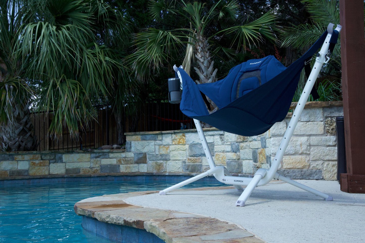 Blue perch hammock chair on a stand by a pool with palm trees in the background