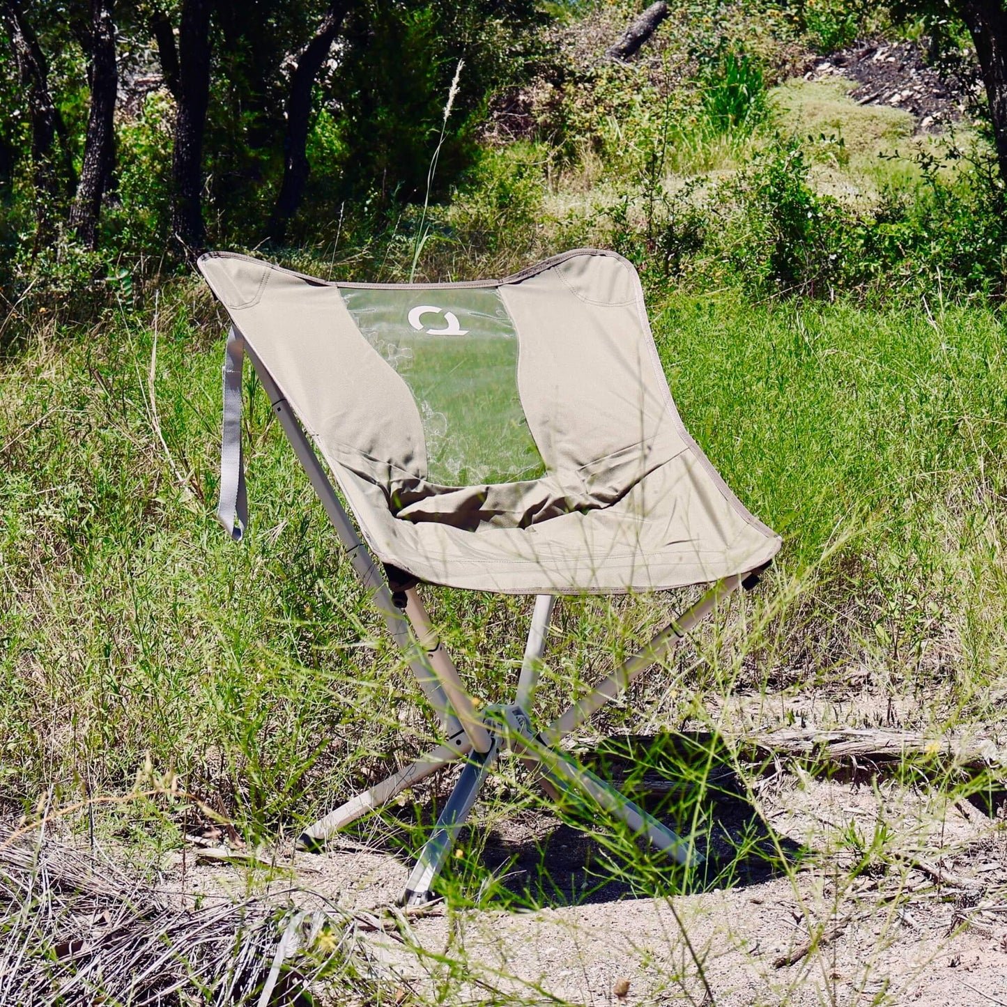 An olive green folding camp chair with a clear back panel sits in a grassy outdoor wooded area. Limited Edition MystiQ Olive Green- CLIQ