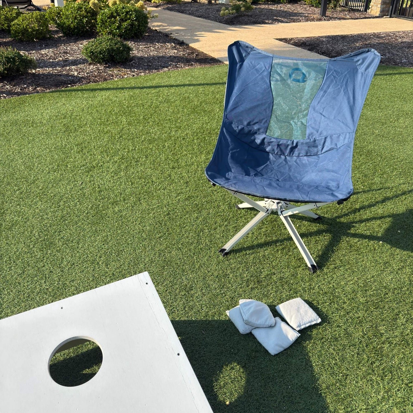 A blue folding patio chair with a white metal frame sits on green artificial turf outdoors, alongside a white bean bag game board and white bean bags. Limited Edition MystiQ Blue - CLIQ