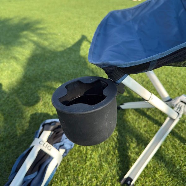 A black cup holder mounted on the arm of a blue and white folding camp chair on green grass outdoors. Limited Edition MystiQ Blue - CLIQ